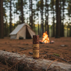Om Naturale Once Bitten Plantain Salve on a wooden camping table with a tent and campfire in the background.